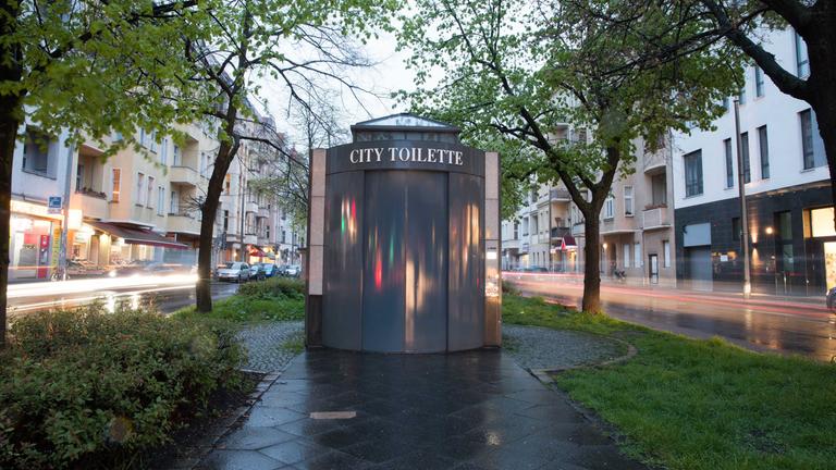 Eine öffentliche Toilette steht auf einem Grünstreifen mit Gehweg zwischen zwei Straßen.