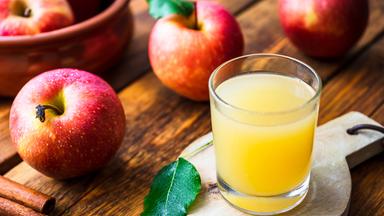 Ein Glas mt naturtrübem Apfelsaft steht af einem Brettchen, drumherum liegen Äpfel.