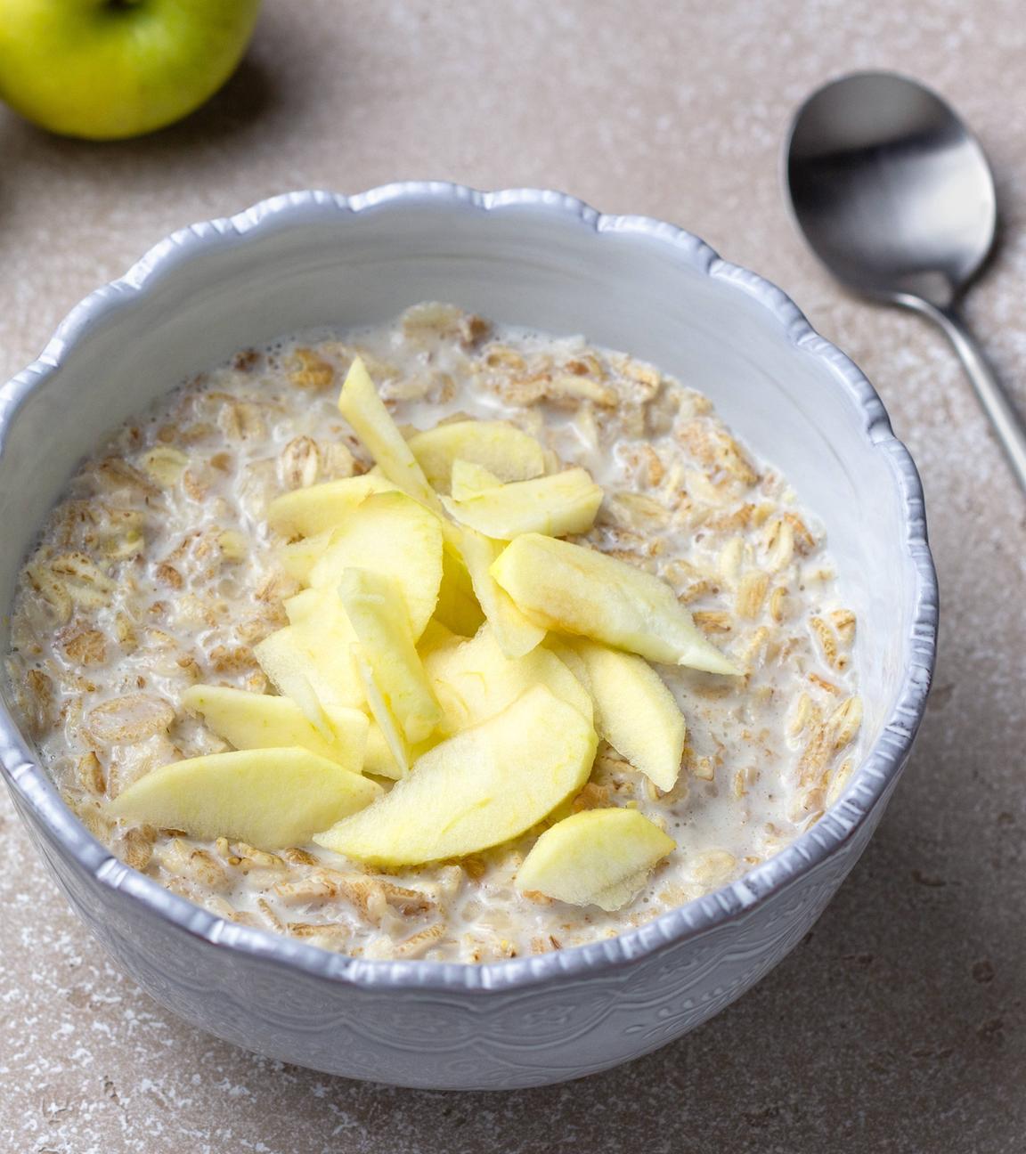 Eine Schale gefüllt mit Porridge, auf dem Apfelschnitze liegen, steht auf einem Tisch, ein Löffel liegt daneben.