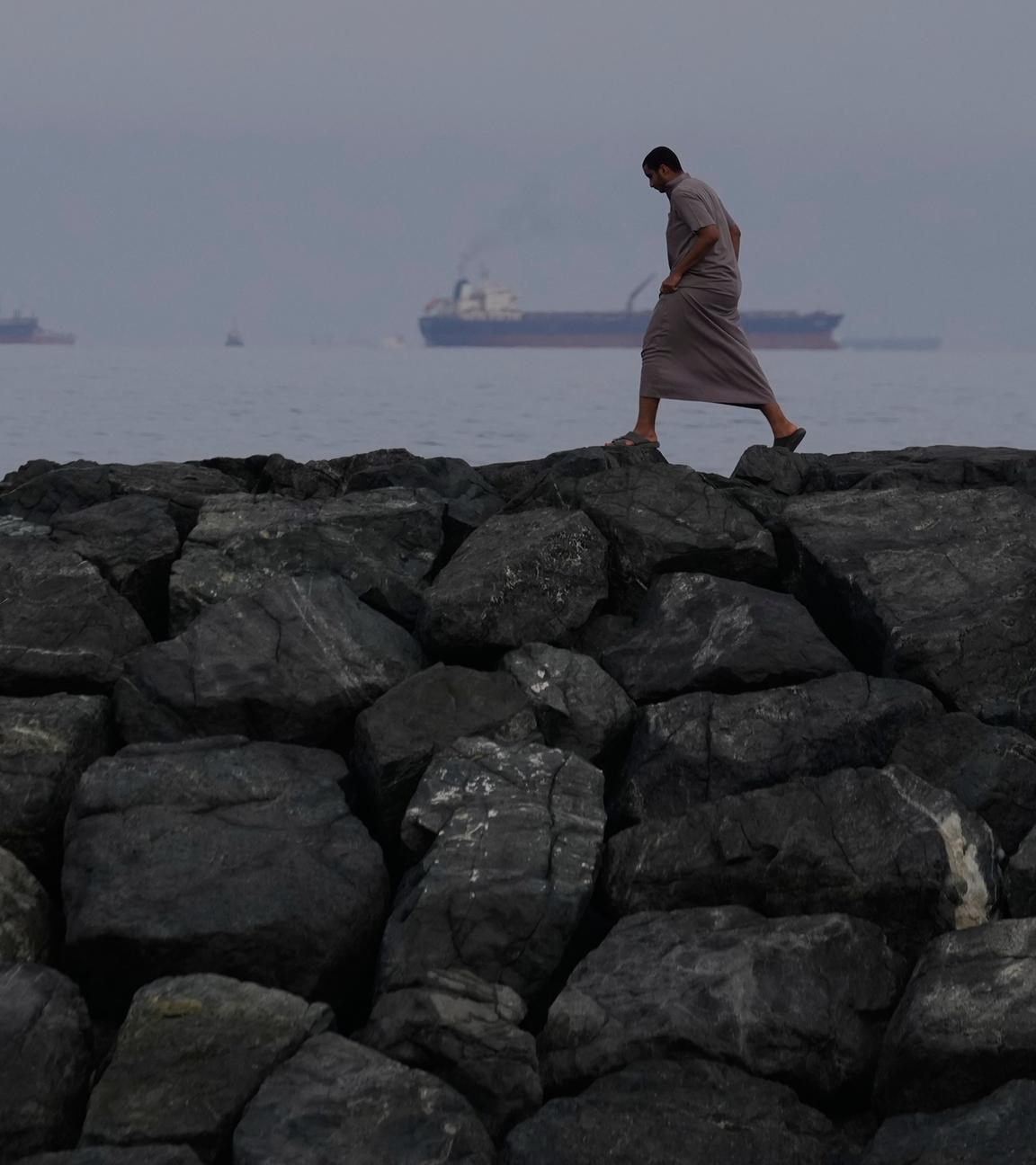 Ein Mann geht auf den Felsen am Ufer spazieren, während Öltanker und Frachtschiffe in der Straße von Hormus,  Vereinigte Arabische Emirate, aufgereiht sind.