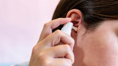 Eine junge Frau hält sich ein Ohrenspray ans Ohr.