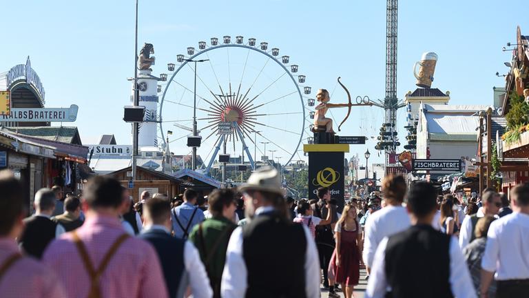 Besucher strömen über das Oktoberfest, im Hintergrund ist ein Riesenrad zu sehen.