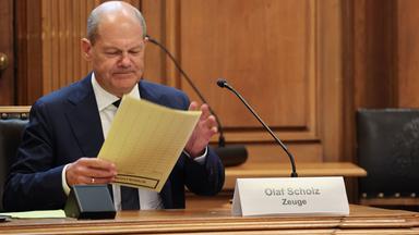 Bundeskanzler Olaf Scholz (SPD) sitzt auf seinem Platz auf der Senatsbank mit einem Plastikschild mit dem Text ·Olaf Scholz Zeuge· vor Beginn einer Sitzung des Parlamentarischen Untersuchungsausschusses ·Cum-Ex· im Plenarsaal der Hamburgischen Bürgerschaft im Rathaus. 