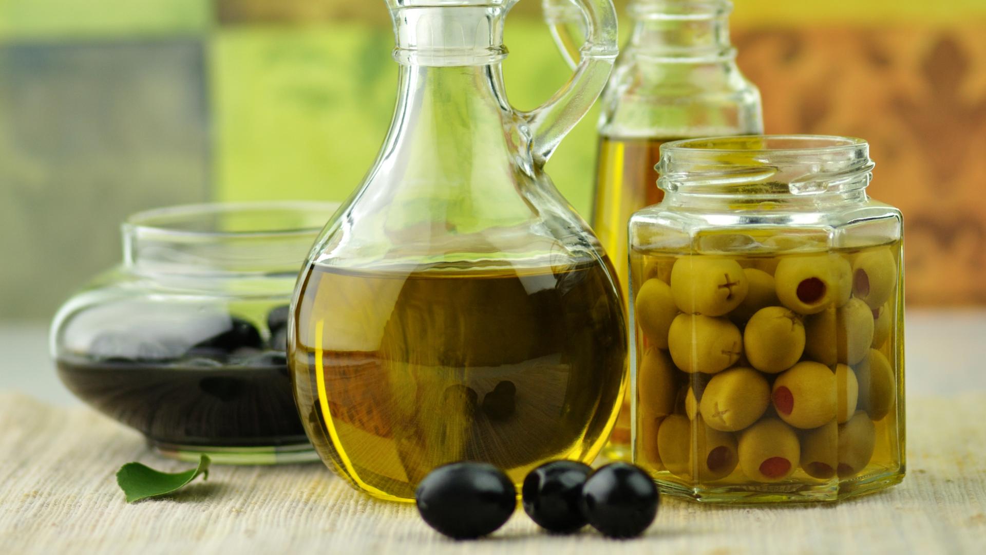 Olivenöl in einer Karaffe steht in der Mitte. Daneben steht ein Einmachglas mit Oliven und weitere Glasgefäße.