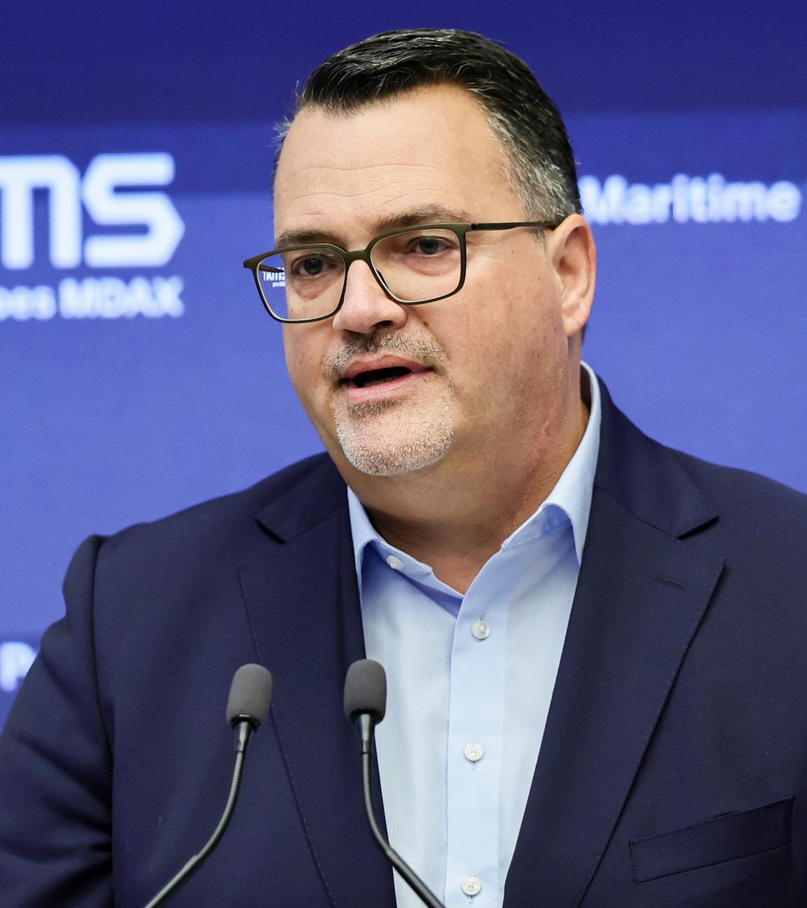 Zu sehen ist Oliver Burkhard, wie er in ein Mikrofon spricht. Er trägt einen Anzug. Der Hintergrund ist blau mit dem Schriftzug TKMS.