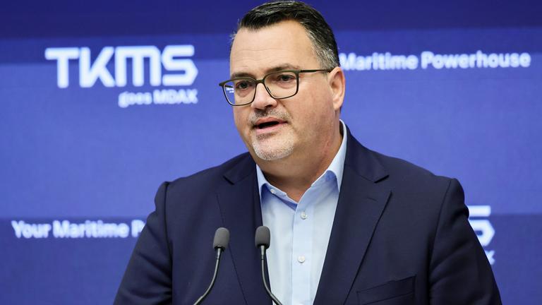 Zu sehen ist Oliver Burkhard, wie er in ein Mikrofon spricht. Er trägt einen Anzug. Der Hintergrund ist blau mit dem Schriftzug TKMS.