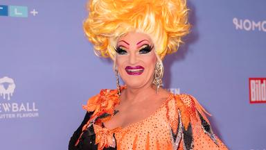 Dragqueen Olivia Jones steht vor einer Fotowand und lächelt in die Kamera. Sie trägt ein orange-schwarzes Kleid und eine blonde Perücke.