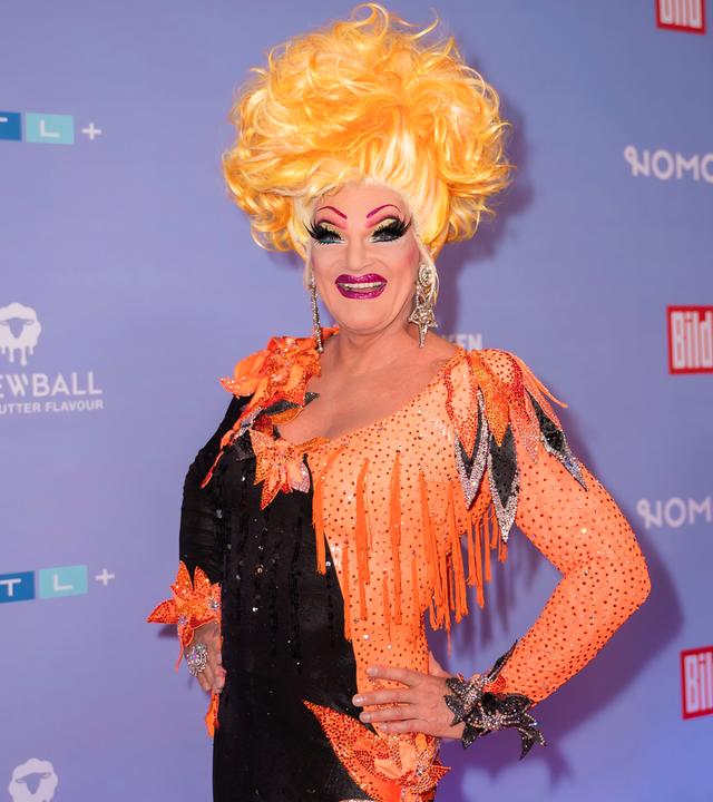 Dragqueen Olivia Jones steht vor einer Fotowand und lächelt in die Kamera. Sie trägt ein orange-schwarzes Kleid und eine blonde Perücke.