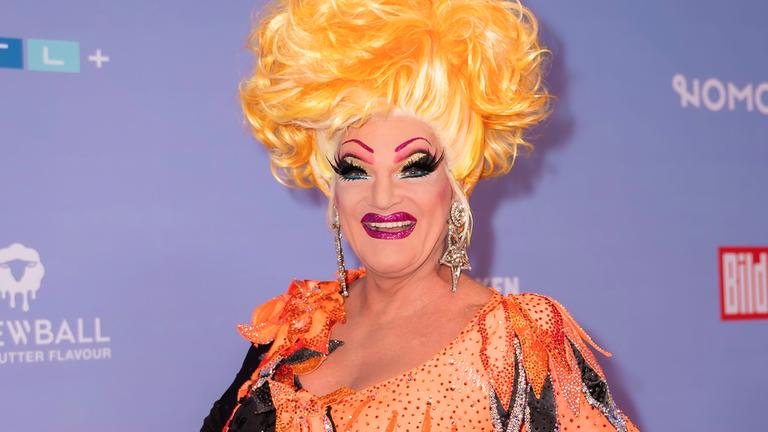 Dragqueen Olivia Jones steht vor einer Fotowand und lächelt in die Kamera. Sie trägt ein orange-schwarzes Kleid und eine blonde Perücke.