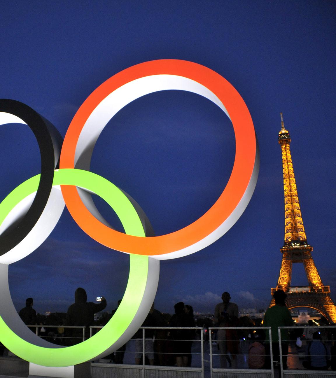 Die beleuchteten olympischen Ringe im Vordergrund. Im Hintergrund der Eiffelturm beleuchtet.