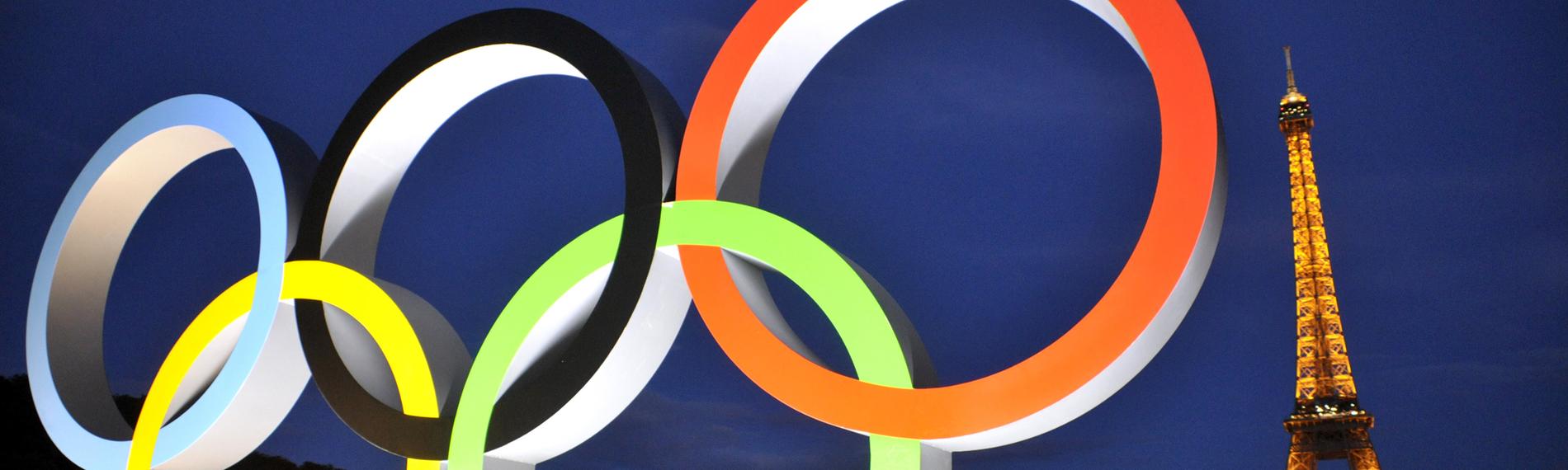 Die beleuchteten olympischen Ringe im Vordergrund. Im Hintergrund der Eiffelturm beleuchtet.