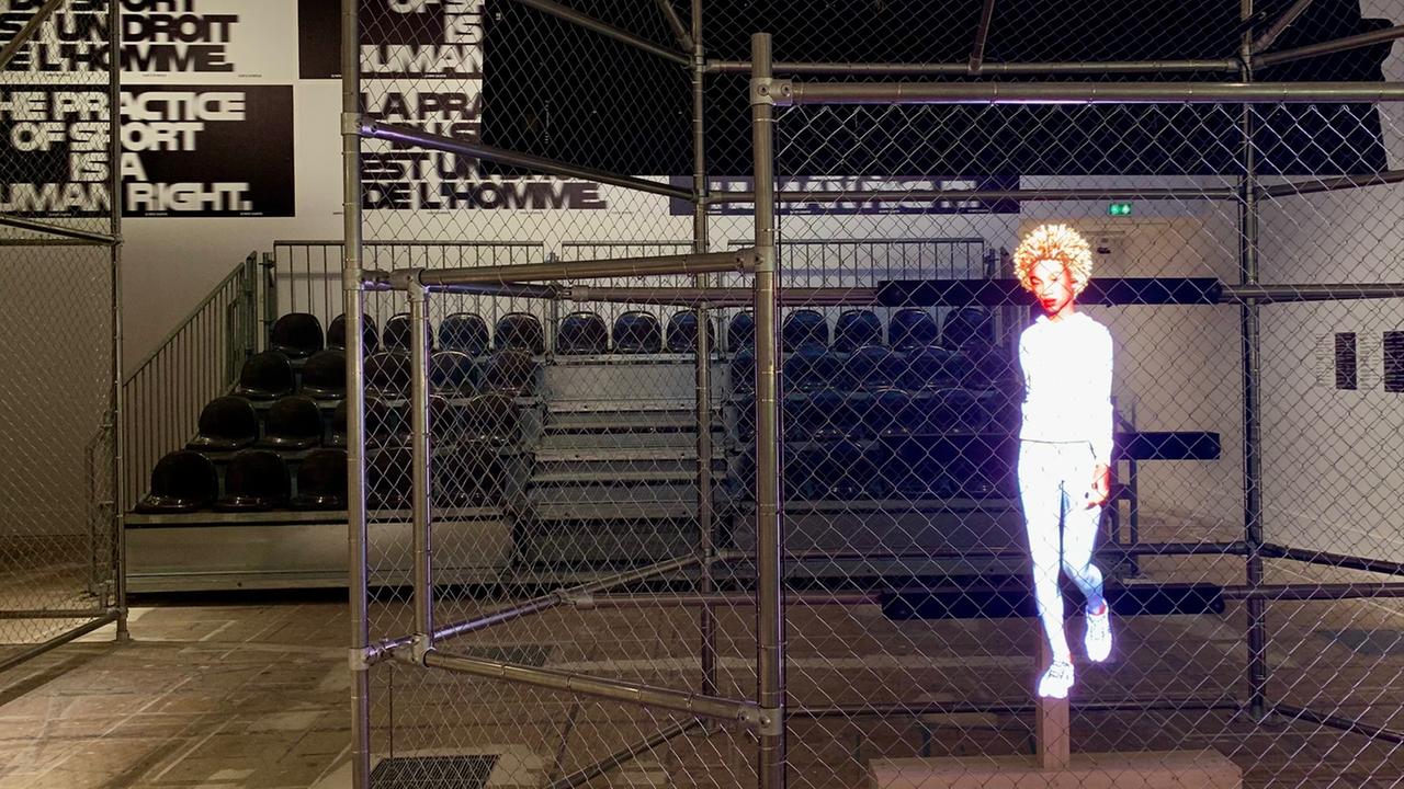 In einer Sporthalle sind Tribüne und Spielfeld mit Metallzäunen voneinander getrennt. Auf die Gitter wird ein Hologramm einer weiß gekleideten, sportlichen Frau projiziert.