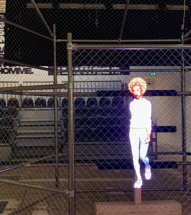 In einer Sporthalle sind Tribüne und Spielfeld mit Metallzäunen voneinander getrennt. Auf die Gitter wird ein Hologramm einer weiß gekleideten, sportlichen Frau projiziert.