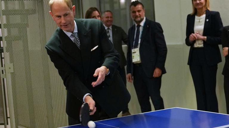 Prinz Edward steht an einer Tischtennis-Platte und schlägt einen Ball auf.