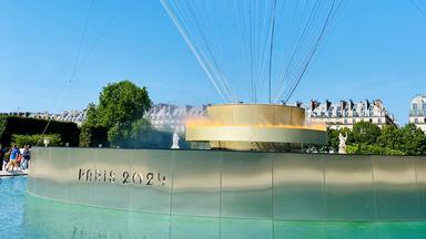In der Mitte eines Wasserbeckens halten kreisförmige Flammen einen goldenen Heißluftballon empor.