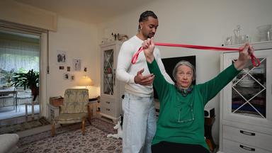 Eva sitzt im Wohnzimmer auf einem Stuhl, hat die Arme nach oben gestreckt und hält ein Fitnessband in den Händen. Louis steht hinter ihr.