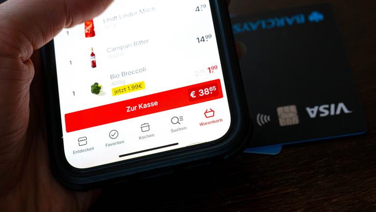 Eine Kreditkarte liegt neben einem Smartphone, auf dem eine App für den Einkauf von Lebensmitteln geöffnet ist.
