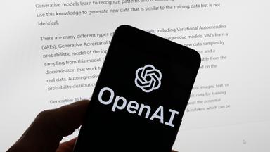 Das Logo von OpenAI auf einem Smartphone. Im Hintergrund ist der Bildschirm eines Computers zu sehen. Darauf ist ein KI-generierter Text zu sehen.
