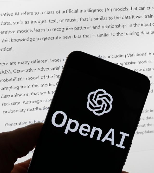 Das Logo von OpenAI auf einem Smartphone. Im Hintergrund ist der Bildschirm eines Computers zu sehen. Darauf ist ein KI-generierter Text zu sehen.
