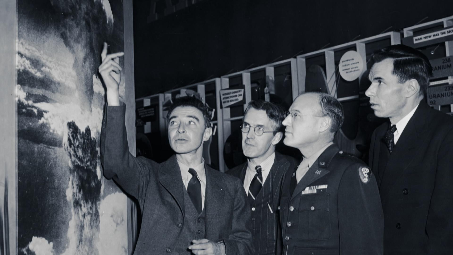 Schwarz-Weiß-Foto: Oppenheimer steht mit drei Regierungsvertretern in einem dunklen Raum vor technischem Gerät. Er deutet auf ein Bild an der Seite, das eine starke Explosion zeigt. Die Regierungsvertreter betrachten es genau.