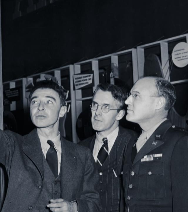 Schwarz-Weiß-Foto: Oppenheimer steht mit drei Regierungsvertretern in einem dunklen Raum vor technischem Gerät. Er deutet auf ein Bild an der Seite, das eine starke Explosion zeigt. Die Regierungsvertreter betrachten es genau.