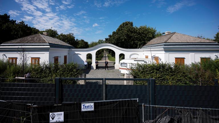 Wachmänner stehen vor einem großen verschlossenen weißen Torbogen. Vor ihnen ist die Zufahrt mit schwarzen Zäunen blockiert.