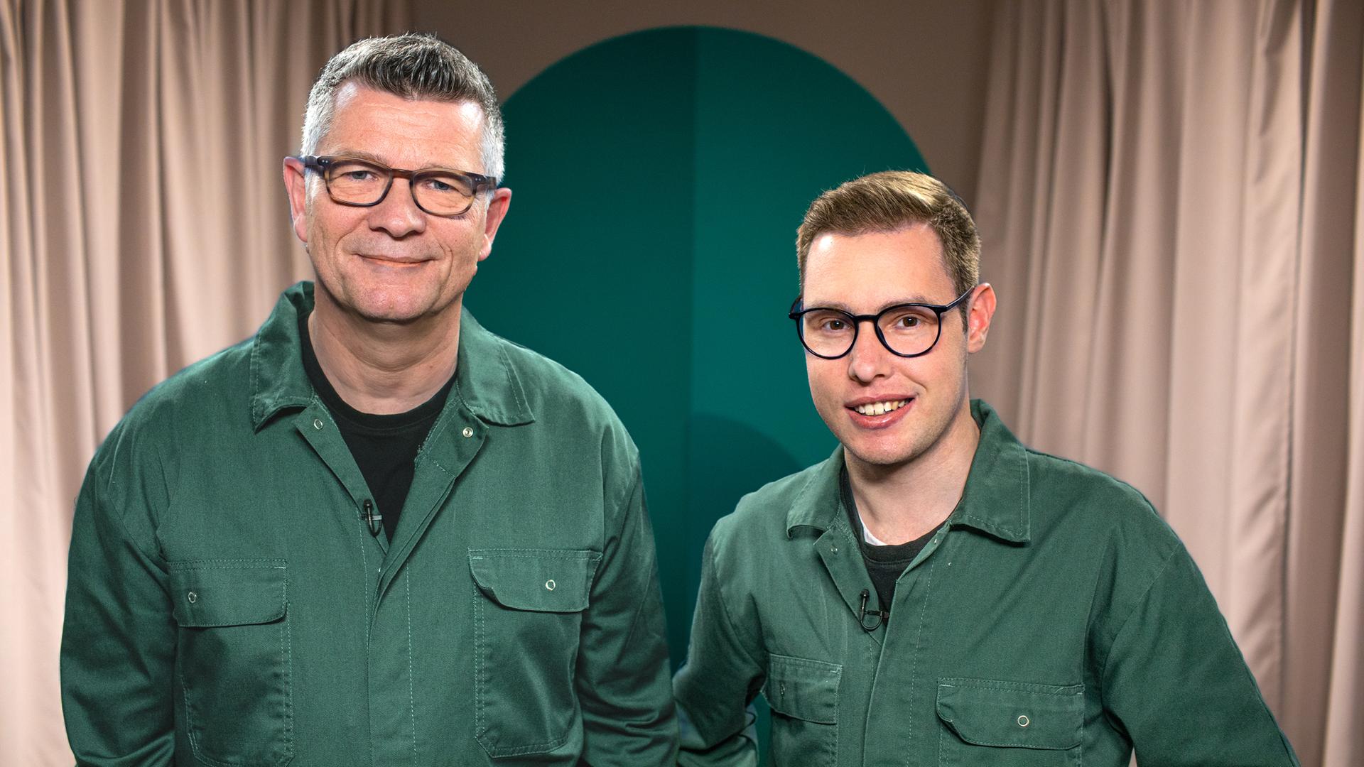 Zwei Menschen in grünen Overalls blicken in die Kamera.