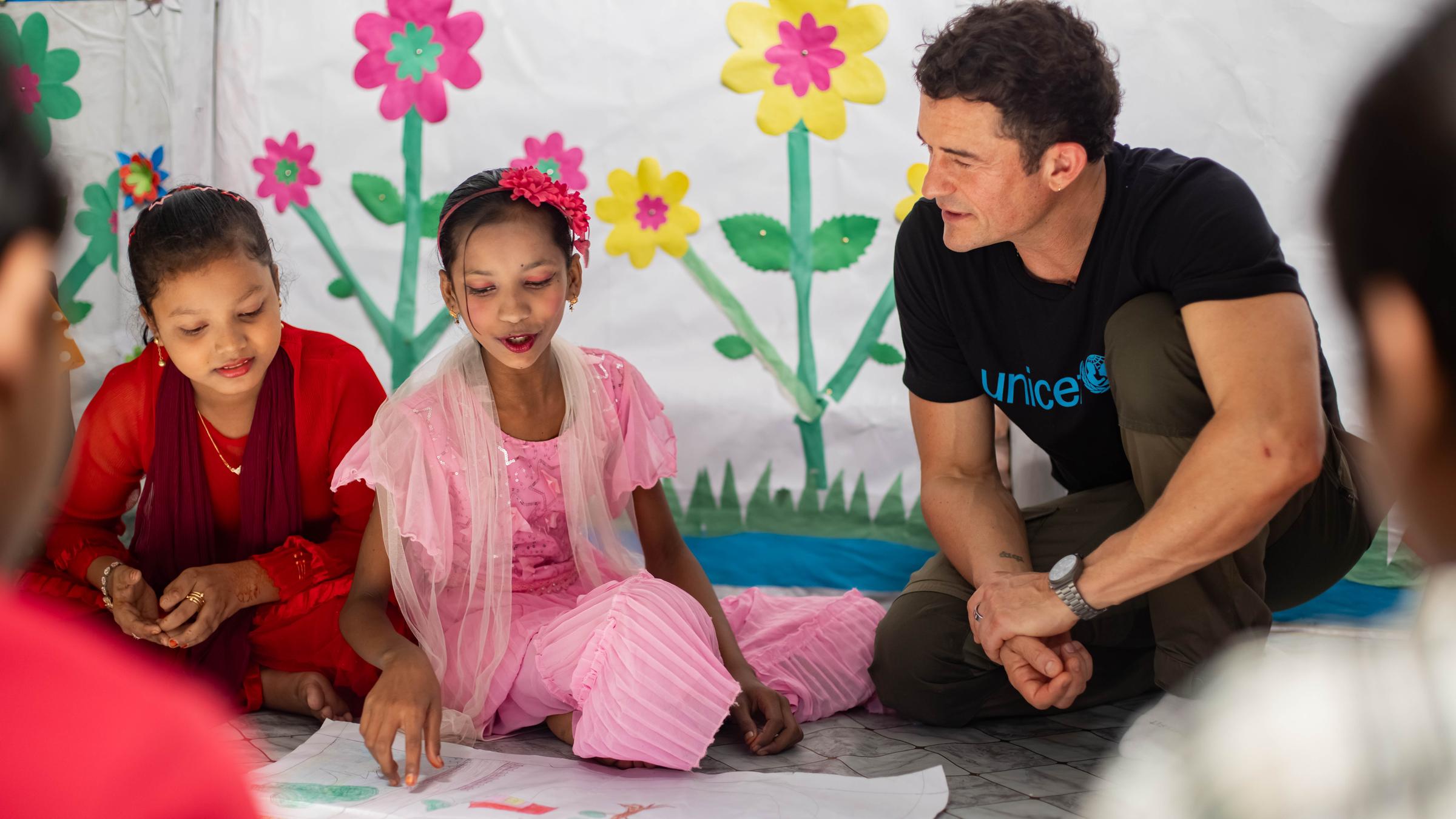Orlando Bloom sitzt mit zwei Mädchen auf dem Boden und lässt sich von einem der Mädchen ein gemaltes Bild vorstellen. 