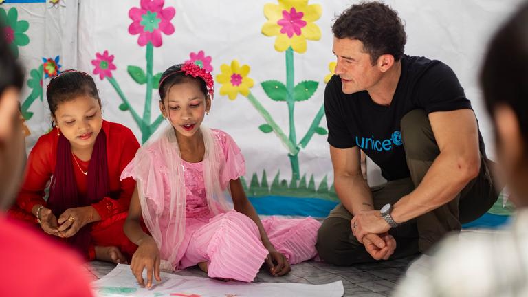 Orlando Bloom sitzt mit zwei Mädchen auf dem Boden und lässt sich von einem der Mädchen ein gemaltes Bild vorstellen. 