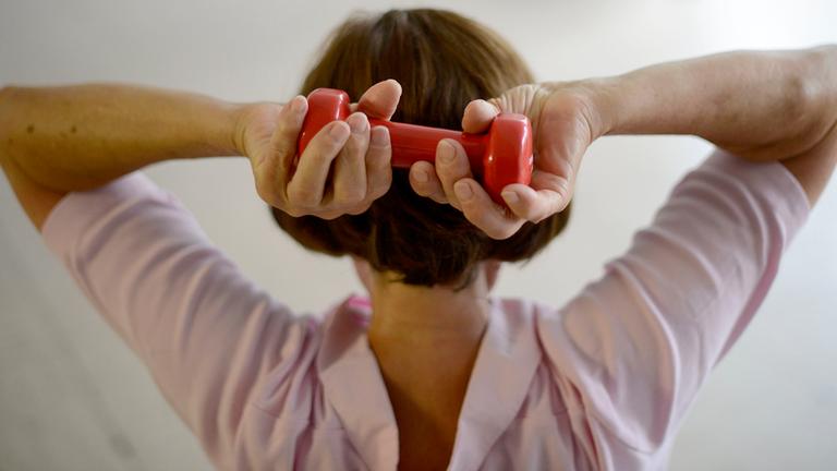 Ältere Frau hält zur Osteoperose-Gymnastik eine kleine Hantel mit beiden Händen hinter dem Kopf. 