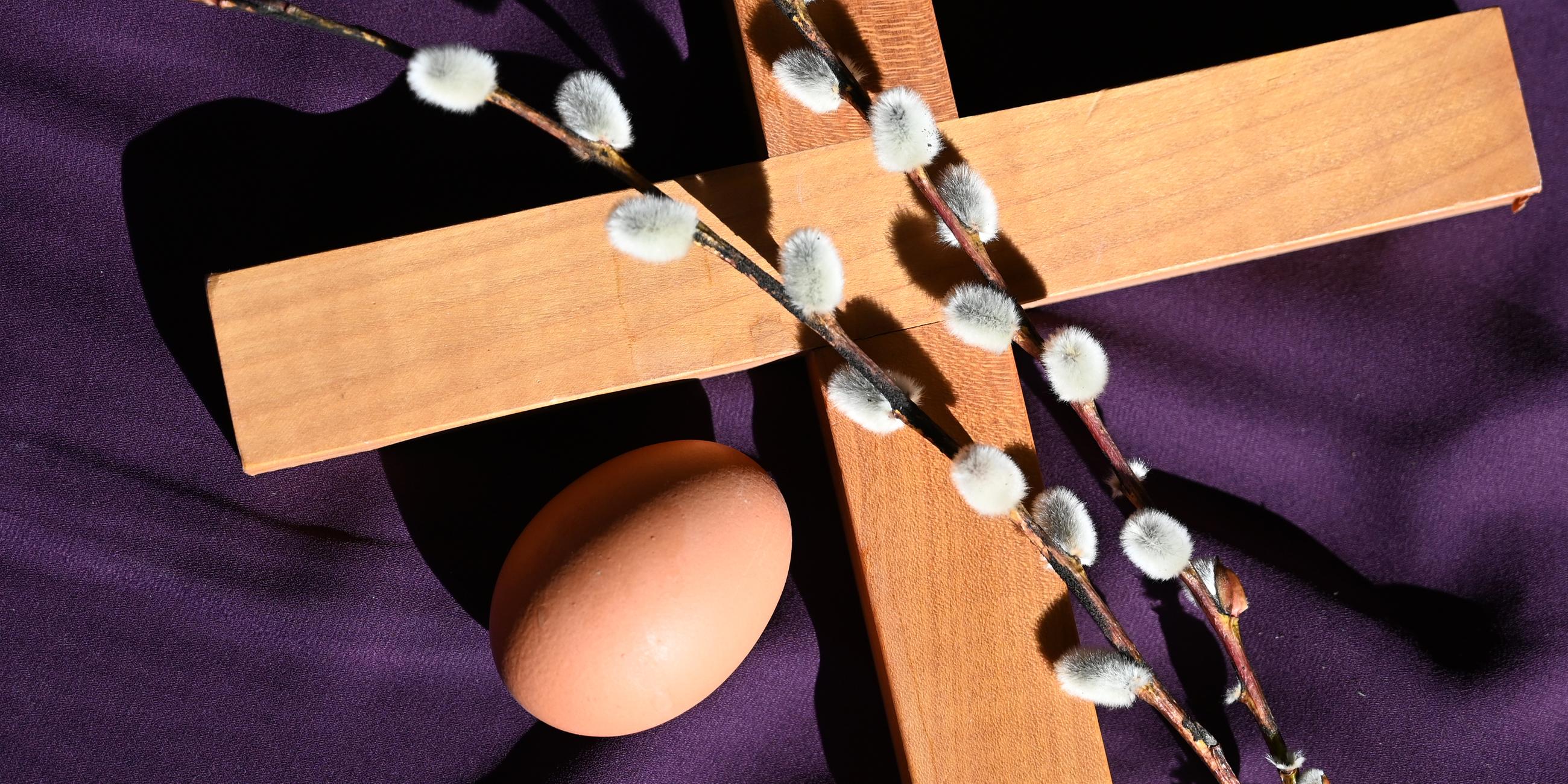 Auf einem lilafarbenen Untergrund liegen ein Kruzifix aus Holz, ein Weidenzweig und ein Hühner-Ei.