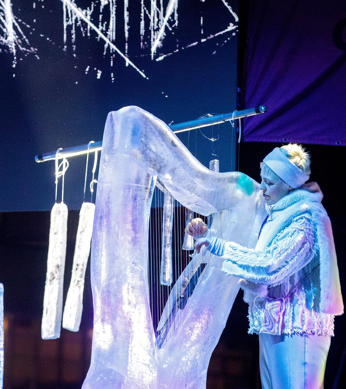 Eine Frau in silbern glitzernder Kleidung spielt eine Harfe aus Eis, im Hintergrund hängt ein Glockenspiel aus Eis.