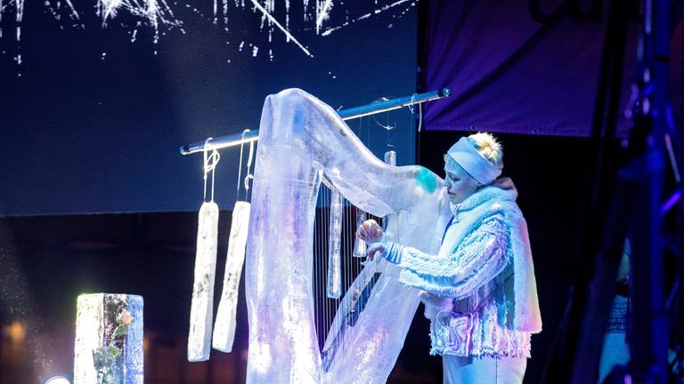 Eine Frau in silbern glitzernder Kleidung spielt eine Harfe aus Eis, im Hintergrund hängt ein Glockenspiel aus Eis.
