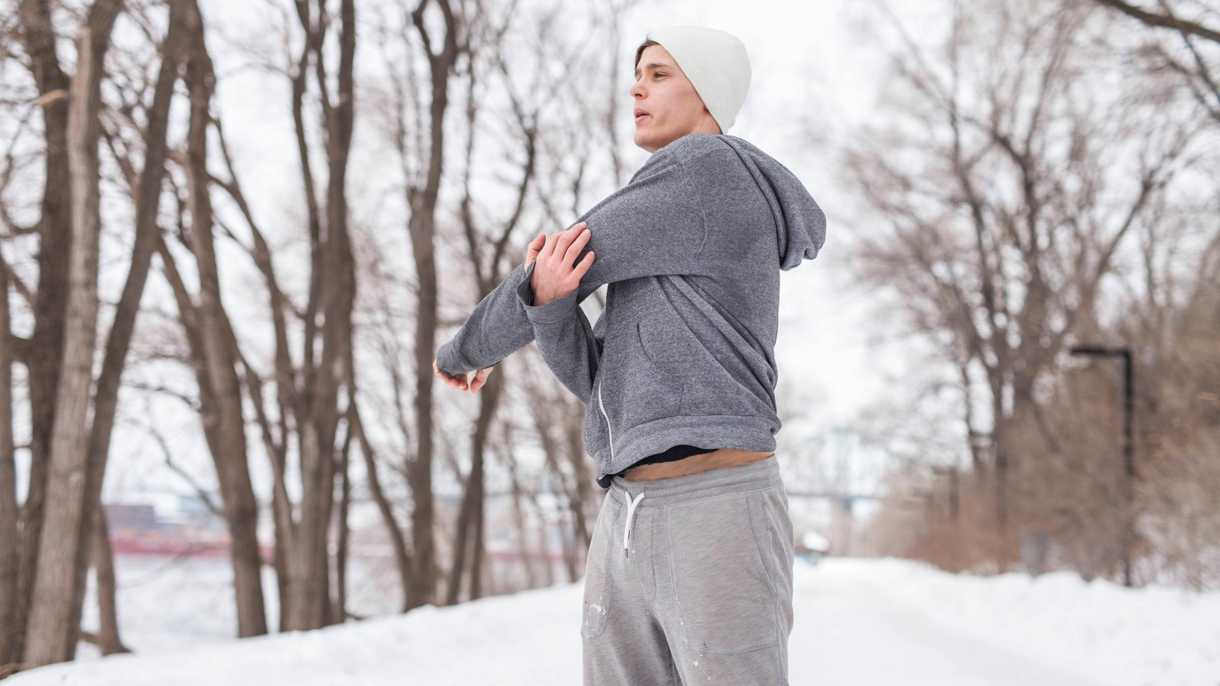 Ein Mann in Trainingskleidung steht in einer verschneiten Landschaft und dehnt sich.