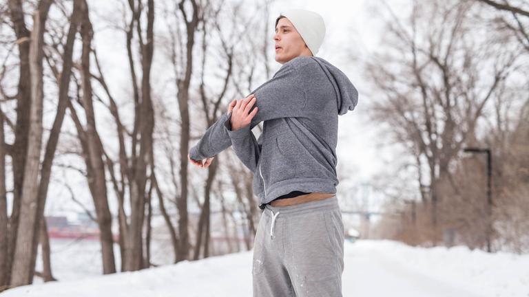 Ein Mann in Trainingskleidung steht in einer verschneiten Landschaft und dehnt sich.