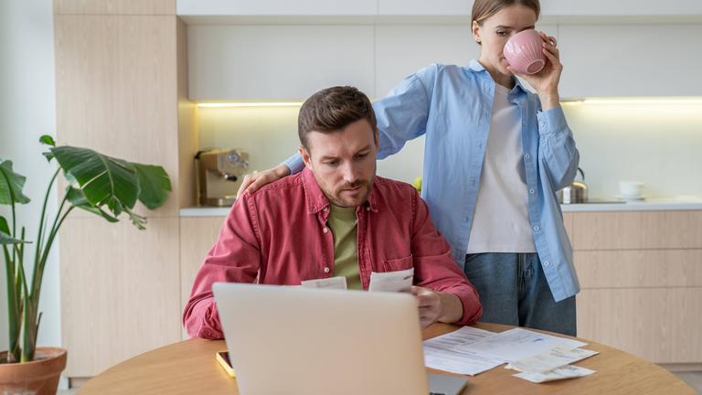 Paar sitzt in der Küche und begutachtet Belege vor einem Laptop.