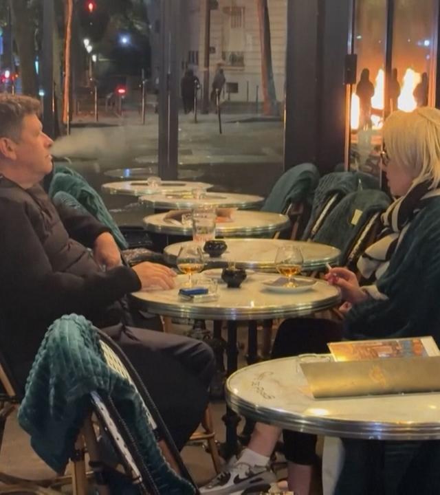 EIin Mann und eine Frau sitzen im Außenbereich eines Lokals in Paris. Im HIntergrund sind ein kleinerer Brand und Einsatzkräfte.