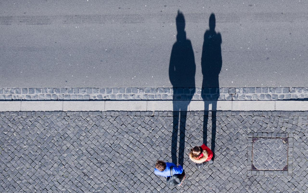 Symbolbild:  Ein Paar steht an einer Straße und wirft bei Sonnenschein lange Schatten auf das Pflaster.