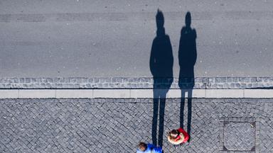 Symbolbild:  Ein Paar steht an einer Straße und wirft bei Sonnenschein lange Schatten auf das Pflaster.