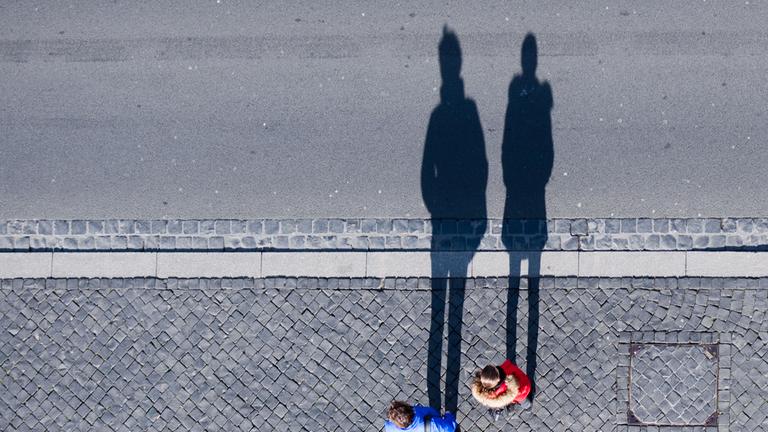 Symbolbild:  Ein Paar steht an einer Straße und wirft bei Sonnenschein lange Schatten auf das Pflaster.