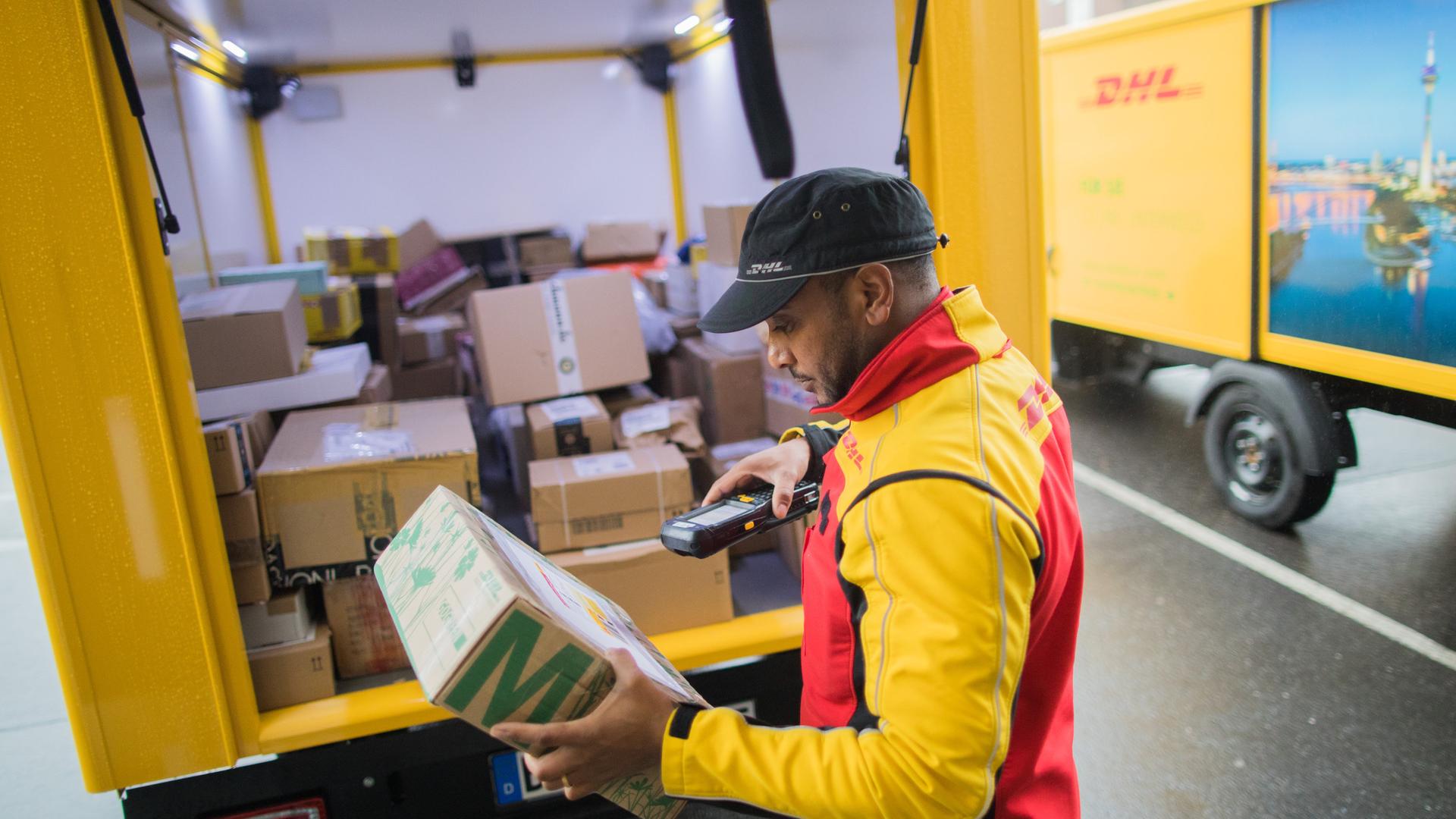 Ein Mitarbeiter steht an einem DHL-Lieferwagen mit geöffneter Ladeklappe und scannt ein Päckchen.