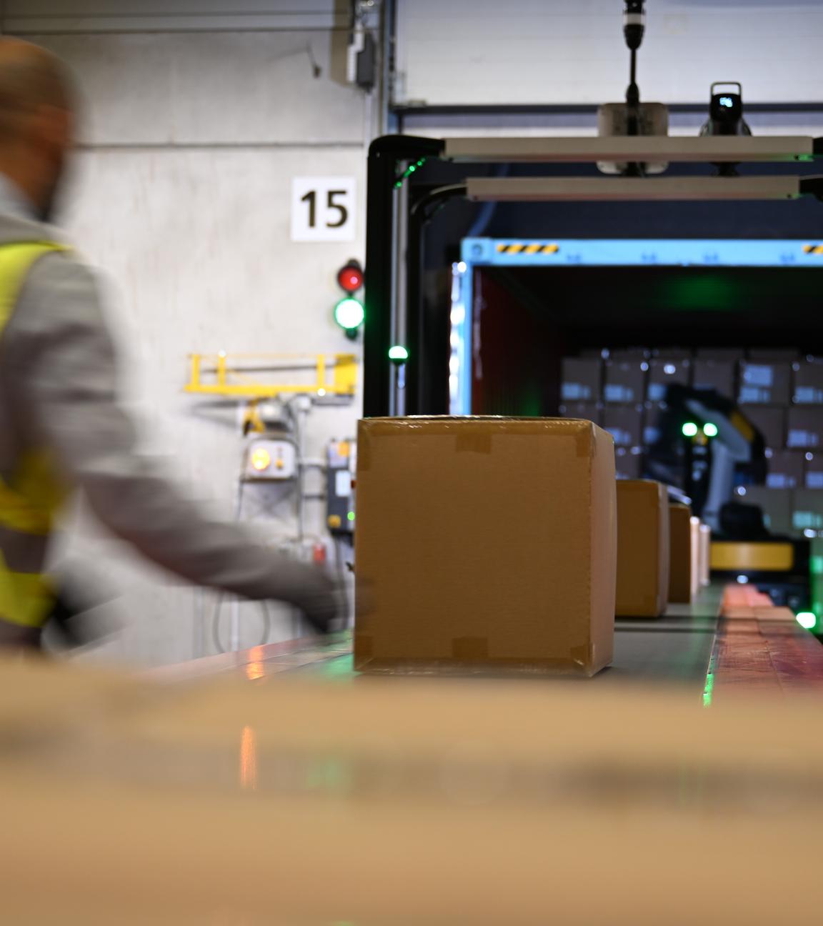 Ein DHL-Mitarbeiter steht in einem Lagerhaus und holt Pakete vom Förderband, die ein Roboter (hinten zu sehen) aus einem Container rausgeholt und auf das Förderband gelegt hat.