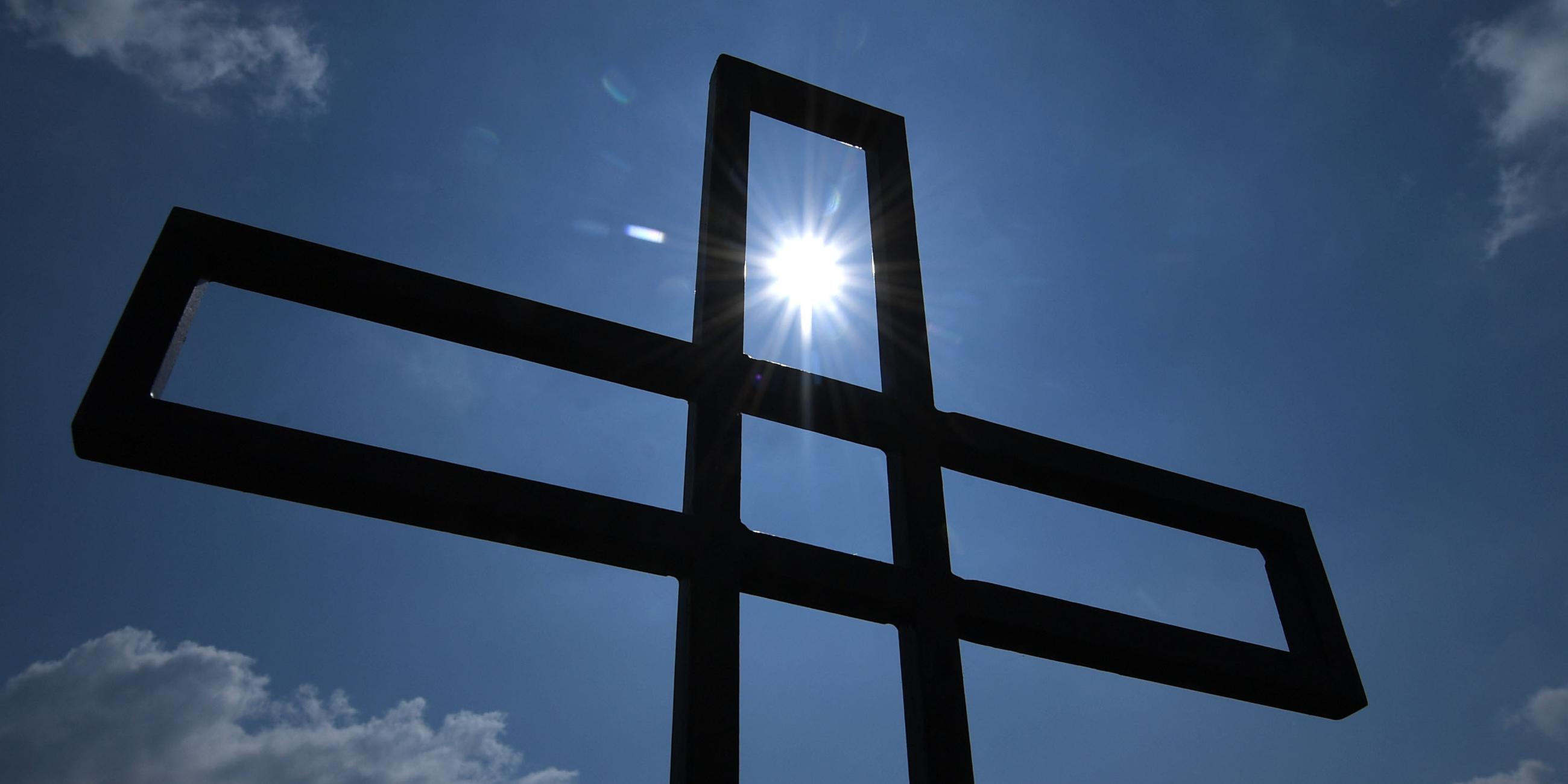 Die Sonne scheint durch ein metallenes Kreuz unter freiem Himmel.