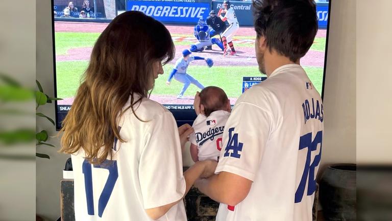Barbara Palvin und Dylan Sprouse stehen in Sportkleidung vor einem Fernseher und halten ihr Baby im Arm. 