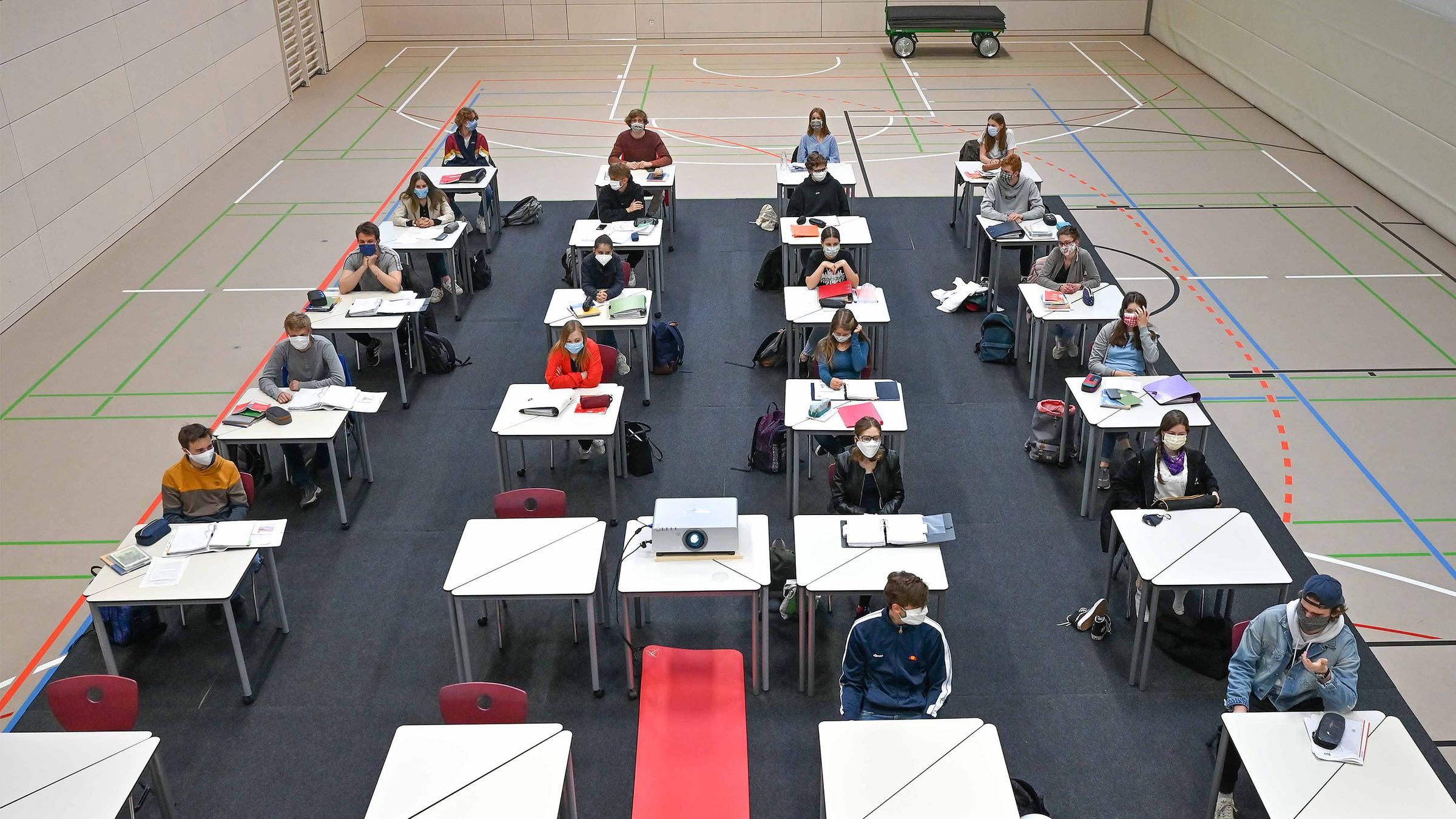 Schüler sitzen an einzelnen Tischen in einer Turnhalle.