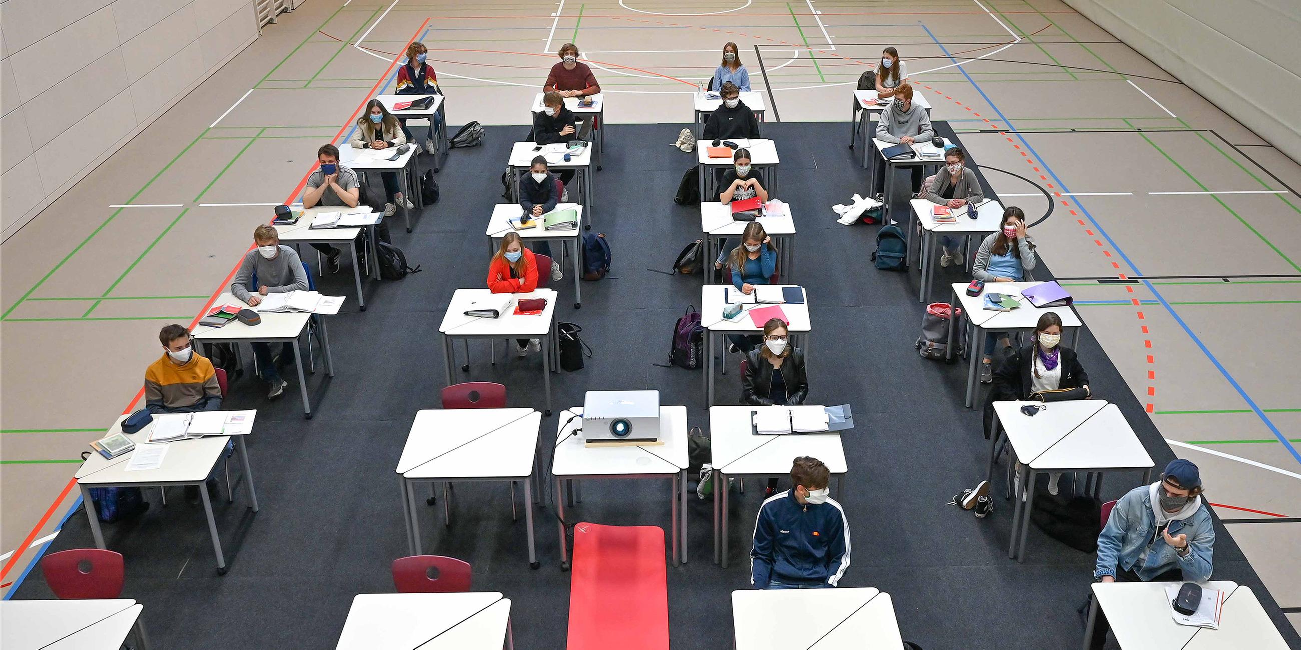 Schüler sitzen an einzelnen Tischen in einer Turnhalle.
