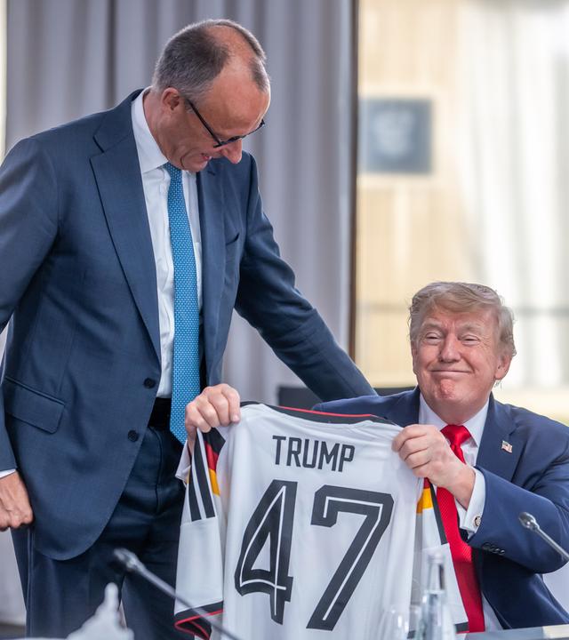 16.06.2026, Frankreich, Evian: Bundeskanzler Friedrich Merz (M, CDU) schenkt ein DFB Fußball Trikot an US-Präsident Donald Trump (r) mit der Rückennummer 47 vor der Arbeitssitzung zu den Themen Welthandel, Iran- und Ukraine-Krieg beim G7 Treffen in Evian während Keir Starmer, Premierminister von Großbritannien, daneben sitzt.