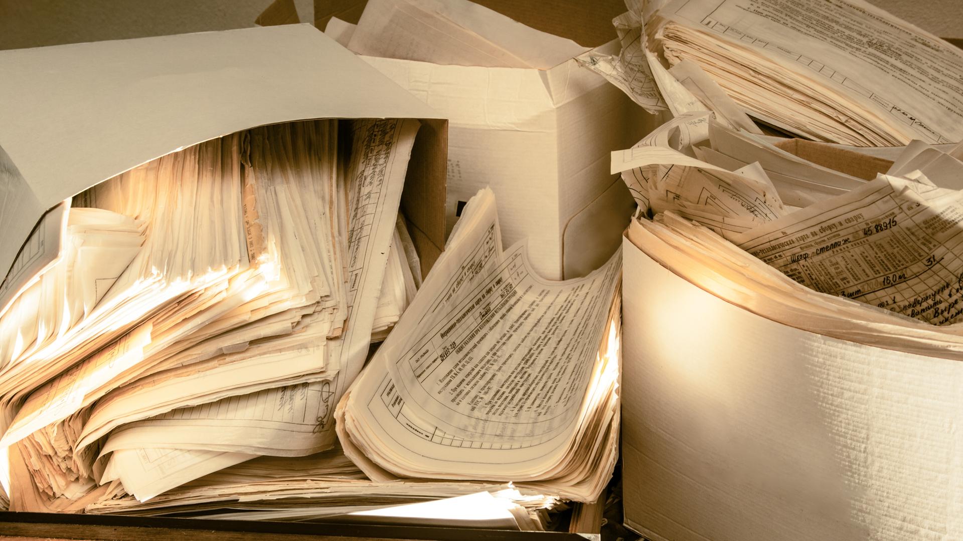 Mehrere Papierstapel teilweise in Kartons liegen durcheinander auf einem Schreibtisch.