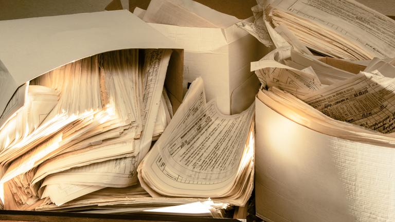 Mehrere Papierstapel teilweise in Kartons liegen durcheinander auf einem Schreibtisch.