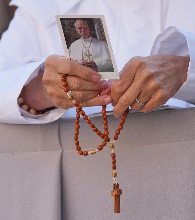 Hände hälten einen roten Rosenkranz und ein Bild von Papst Leo XIV. 